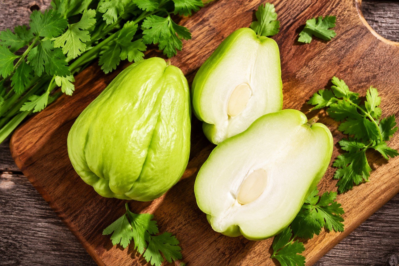Fresh chayote squash — one whole and one sliced in half — resting on a rustic wooden cutting board with sprigs of parsley and cilantro. You can see the pale white flesh and single flat seed inside the cut half. Soft morning light, cozy kitchen vibe.