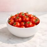 Fresh red cherry tomatoes in a matte white ceramic bowl on a light marble countertop with soft shadows and a pastel background.