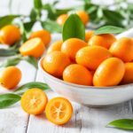 Fresh kumquats with water droplets, some sliced open, on a rustic white wooden table with green leaves in natural light