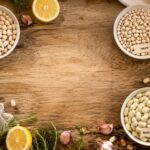 Overhead flat lay of cannellini, navy, Great Northern, and baby lima beans in white ceramic bowls with garlic, lemon, rosemary, olive oil, and leafy greens on a rustic wooden table.