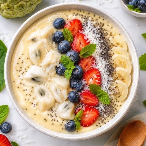Overhead view of a creamy sugar apple smoothie bowl topped with fresh berries, banana slices, chia seeds, coconut flakes, and mint leaves.