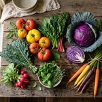 A top-down view of various types of food including fresh anti-inflammatory vegetables like tomatoes, kale, purple cabbage, and rainbow carrots arranged on a rustic wooden table.